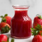 A glass jar of sweet, ruby-red strawberry syrup surrounded by whole fresh strawberries.
