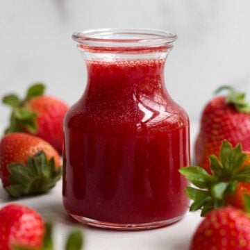 A glass jar of sweet, ruby-red strawberry syrup surrounded by whole fresh strawberries.