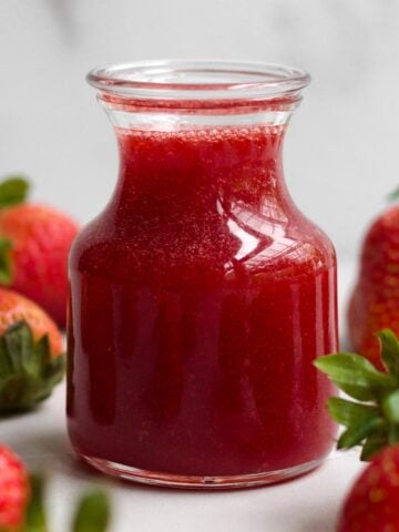 A glass jar of sweet, ruby-red strawberry syrup surrounded by whole fresh strawberries.
