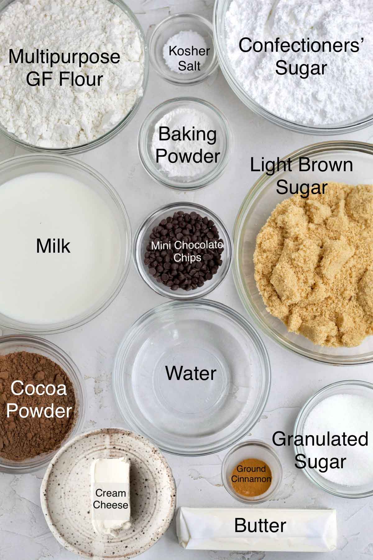 Chocolate cinnamon rolls ingredients in separate bowls: multipurpose gluten free flour, kosher salt, confectioners' sugar, baking powder, milk, mini chocolate chips. light brown sugar, cocoa powder, water, granulated sugar, cream cheese, butter, ground cinnamon and butter.