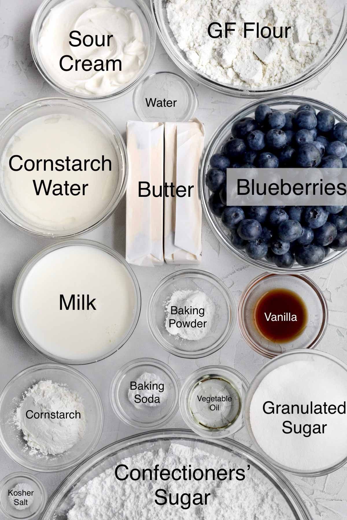 Blueberry cupcakes ingredients in separate bowls: Sour cream, gluten free flour, cornstarch water, water, butter, blueberries, milk, baking powder, vanilla, cornstarch, baking soda, vegetable oil, granulated sugar, kosher salt, and confectioners' sugar.