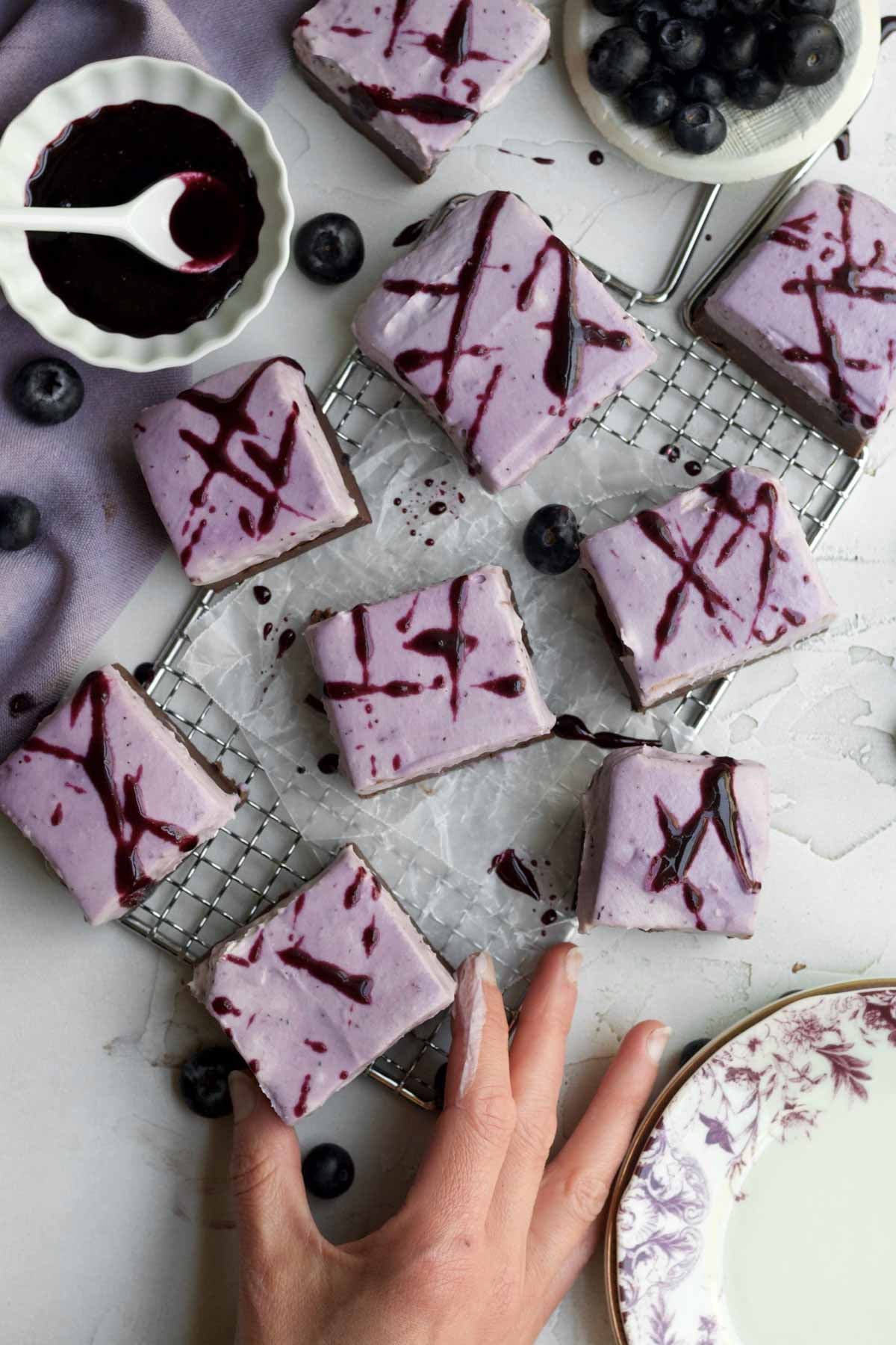 Several creamy blueberry fudge bars squares with lavender blueberry cream cheese topping with a hand grabbing one.
