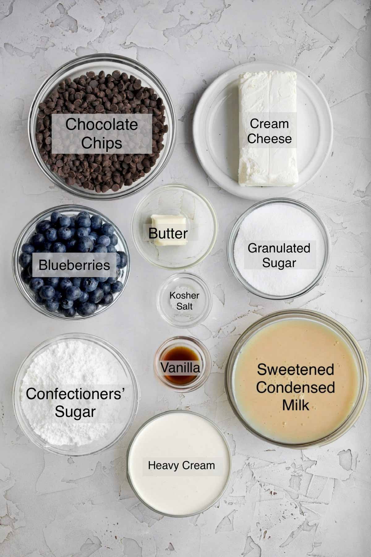 Blueberry fudge bars ingredients in separate bowls: chocolate chips, cream cheese, blueberries, butter, granulated sugar, kosher salt, confectioners' sugar, vanilla, sweetened condensed milk, and heavy cream.