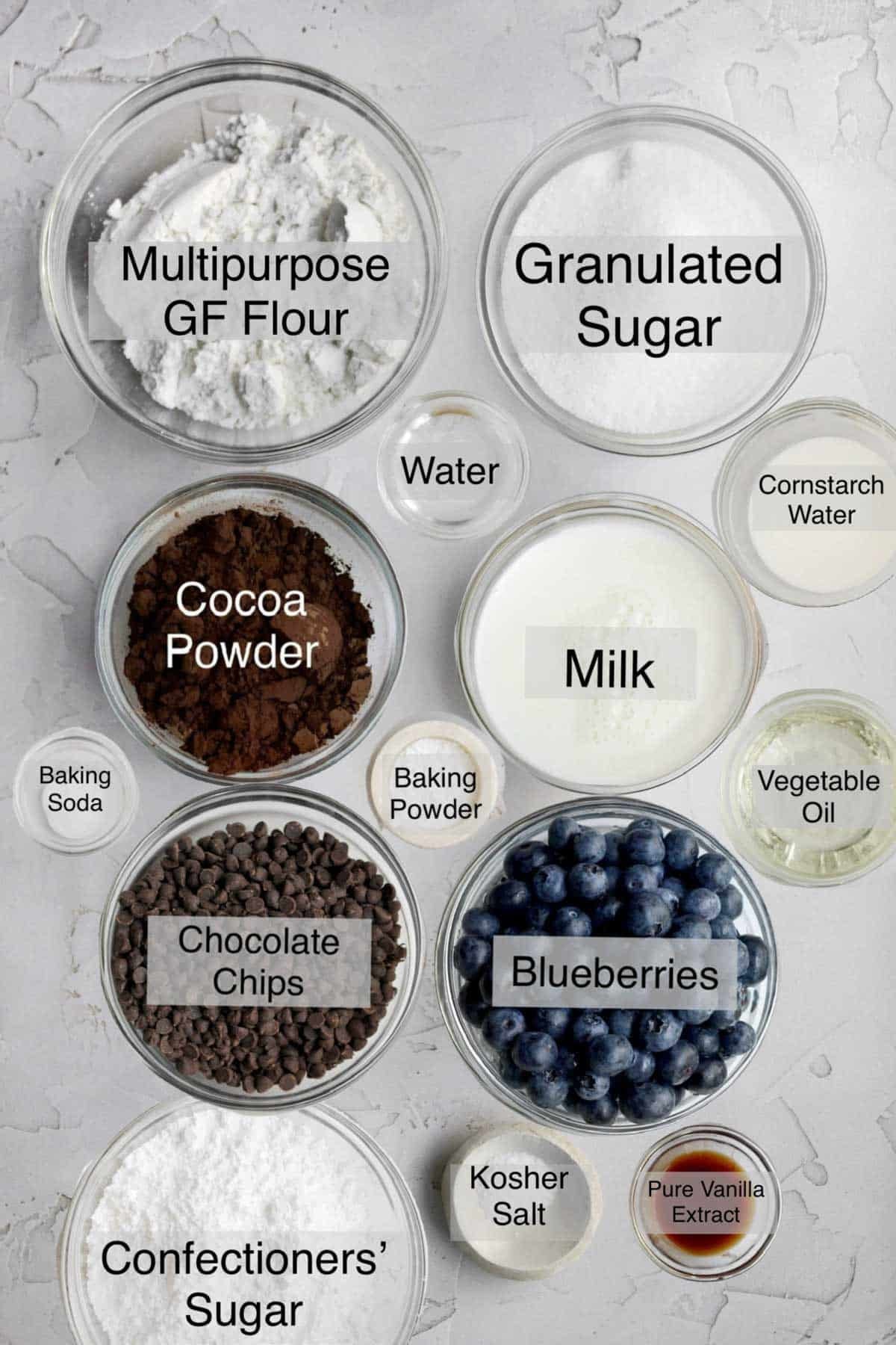 Chocolate blueberry muffins ingredients in separate bowls: multipurpose gluten free flour, granulated sugar, cocoa powder, water, milk, cornstarch water, baking soda, baking powder, vegetable oil, chocolate chips, blueberries, confectioners' sugar, kosher salt and pure vanilla extract.