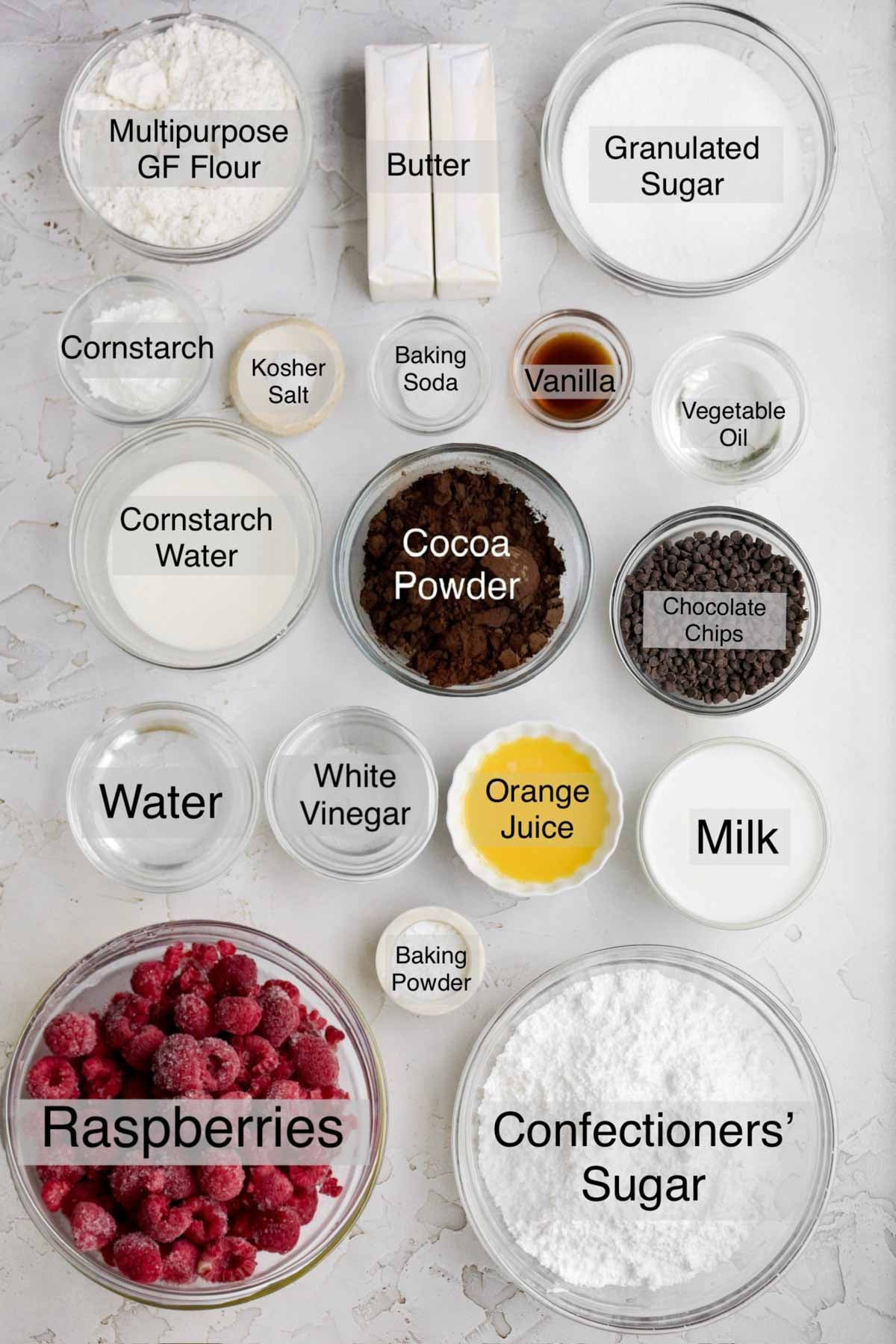 Chocolate raspberry cupcakes ingredients in separate bowls: multipurpose gluten free flour, butter, granulated sugar, cornstarch, kosher salt, baking soda, vanilla, vegetable oil, cornstarch water, cocoa powder, chocolate chips, water, white vinegar, orange juice, milk, baking powder, raspberries and confectioners' sugar.