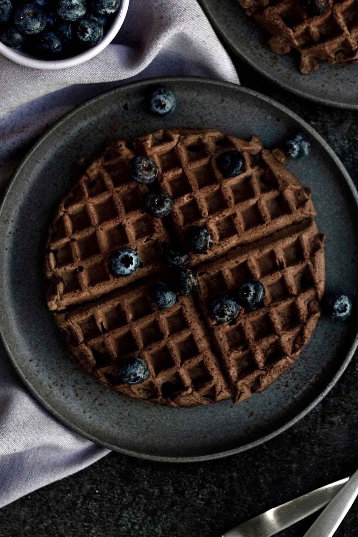 Round uncut chocolate waffles on black plates with blueberries.