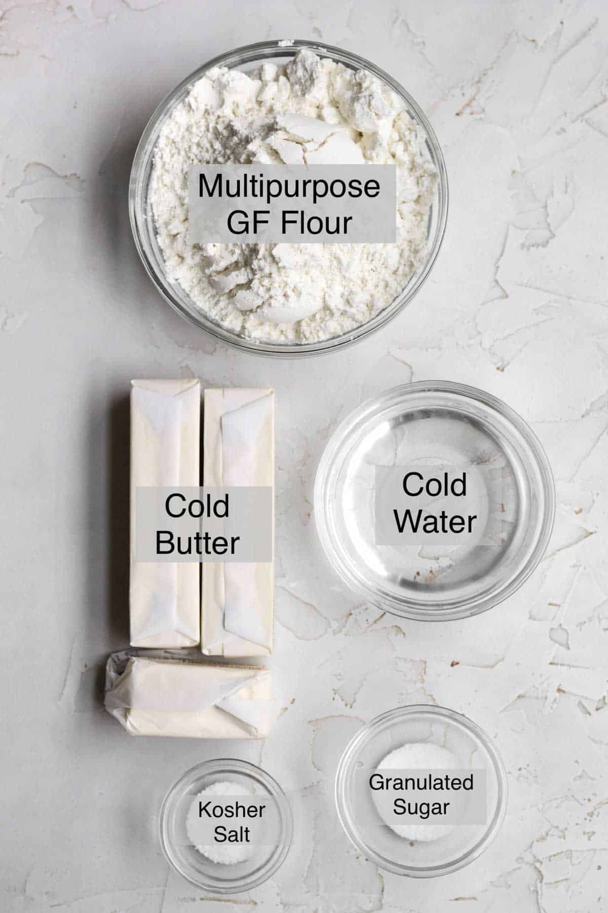 Easy puff pastry ingredients in separate bowls: Multipurpose gluten free flour, cold butter, cold water, kosher salt, and granulated sugar.