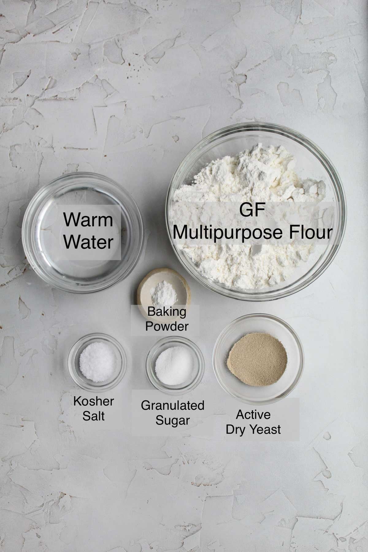 Gluten free pizza dough ingredients in separate containers: warm water, gluten free multipurpose flour, baking powder, kosher salt, granulated sugar, and active dry yeast.