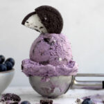 A scoop of purple Oreo blueberry ice cream topped with a halved gluten-free chocolate sandwich cookie.