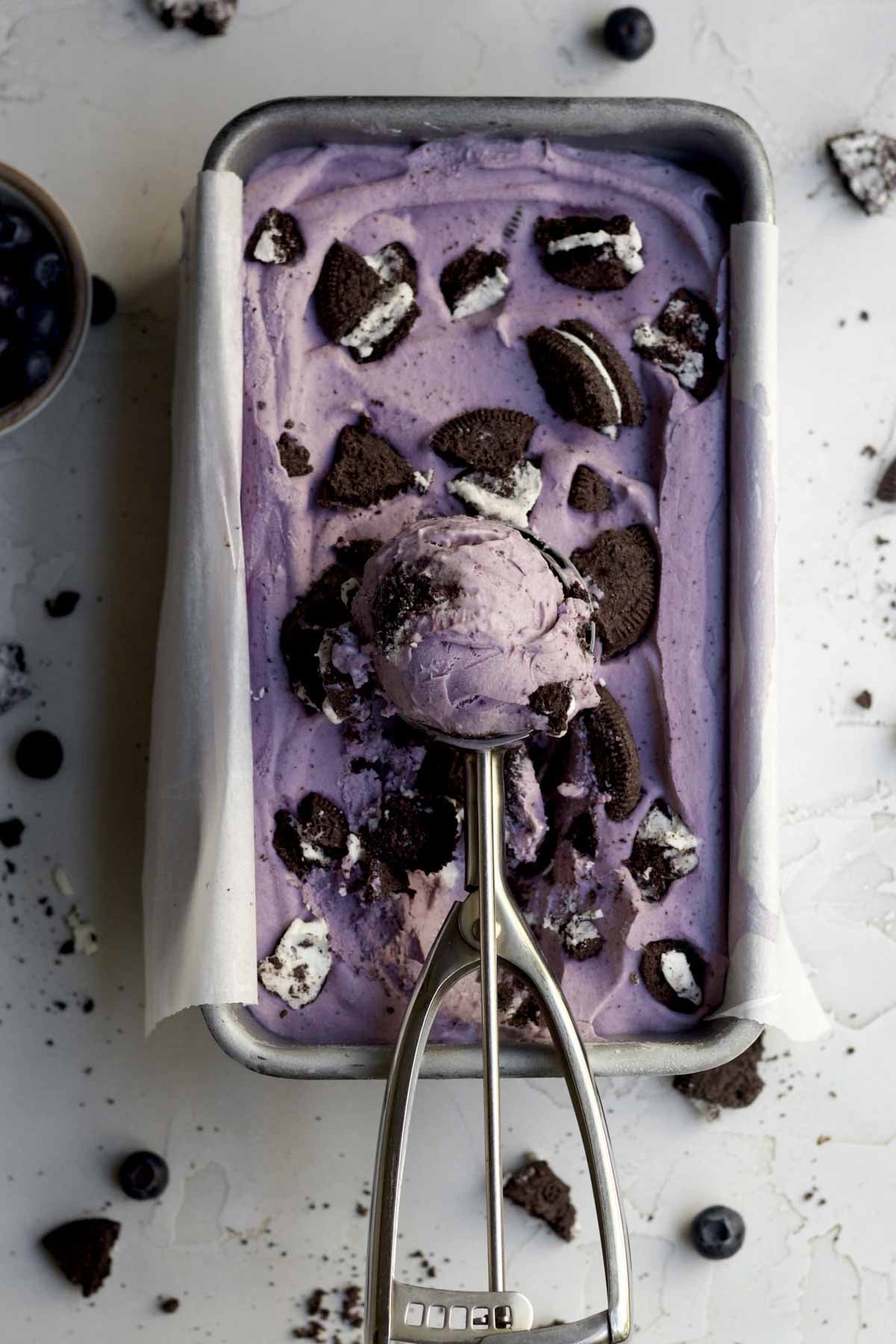 A silver scoop filled with Oreo blueberry ice cream sits atop a metal pan of the same.