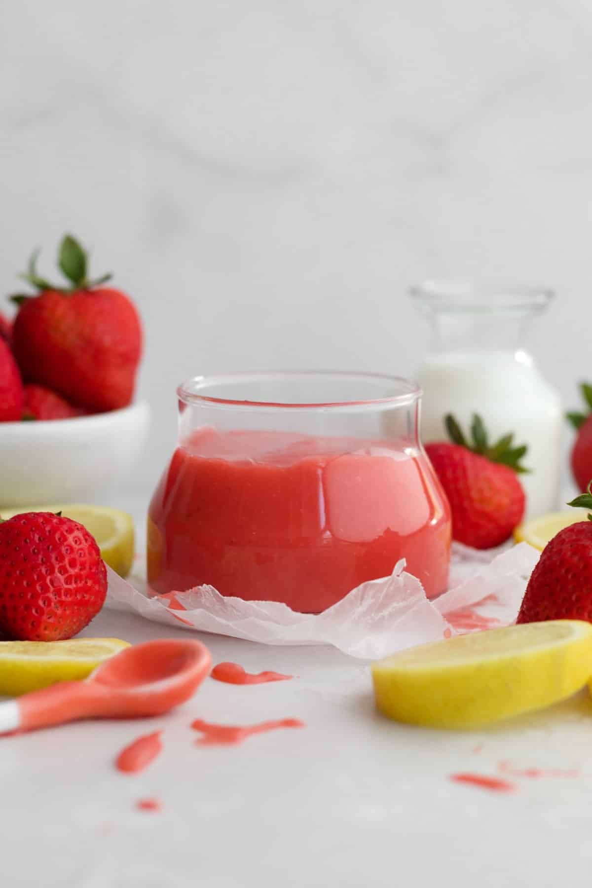 Strawberry curd sits sweetly in a jar amongst fresh strawberries and lemon slices.