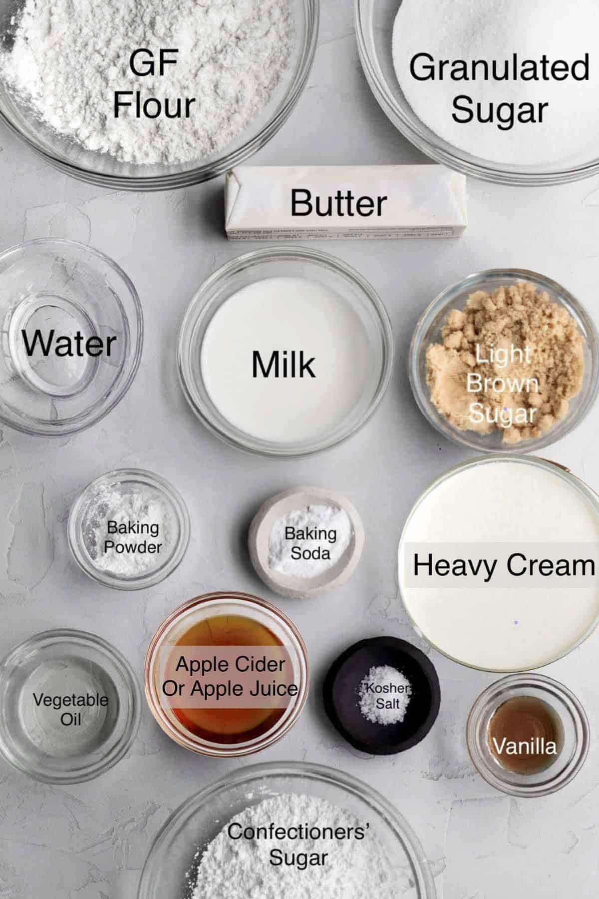 Gluten free apple cake ingredients in separate bowls: Gluten free flour, granulated sugar, butter, water, milk, light brown sugar, baking powder, baking soda, heavy cream, vegetable oil, apple cider or apple juice, kosher salt, vanilla and confectioners' sugar.
