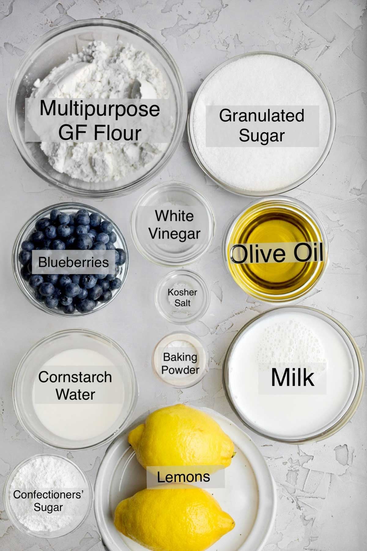 Gluten free olive oil cake ingredients in separate bowls: multipurpose gluten free flour, granulated sugar, blueberries, white vinegar, olive oil, kosher salt, cornstarch water, baking powder, milk, confectioners' sugar and lemons.