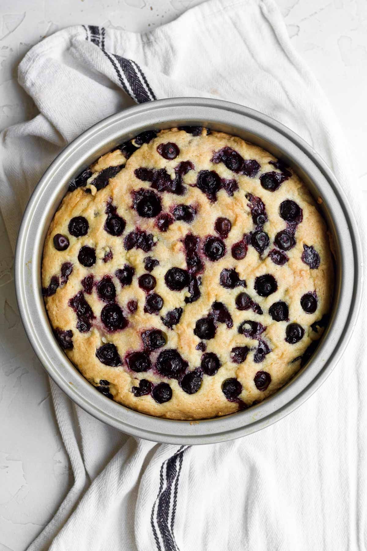 Golden-brown gluten free olive oil cake in a cake pan with warm bursted blueberries.