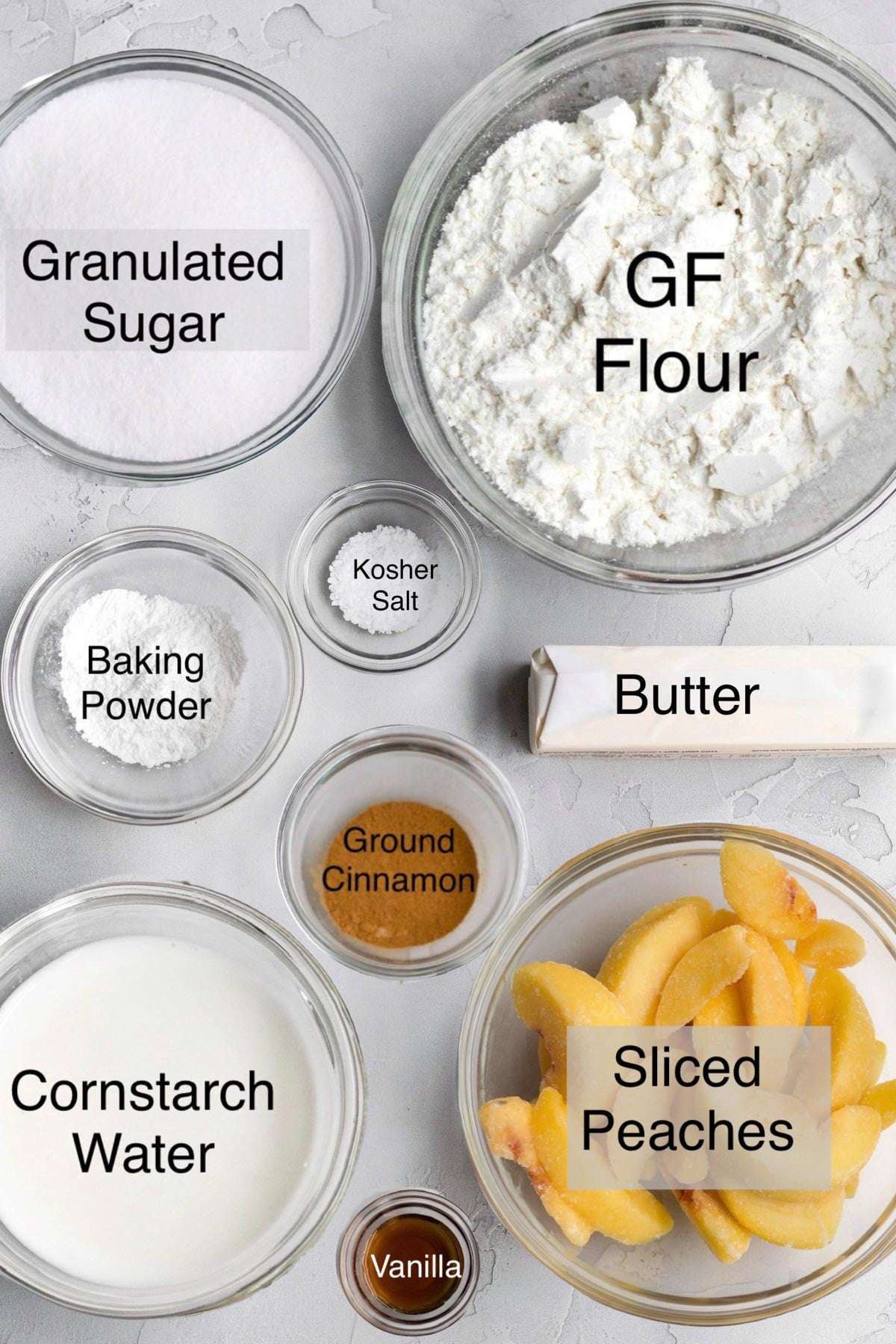Gluten free peach cobbler ingredients in separate bowls: Granulated sugar, gluten free flour, baking powder, kosher salt, butter, ground cinnamon, cornstarch water, vanilla, and sliced peaches.
