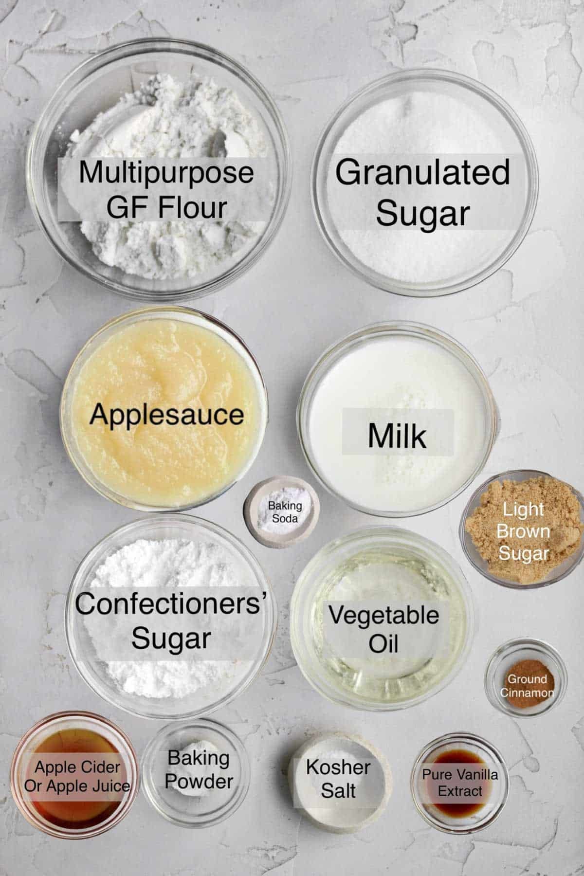 Apple loaf cake ingredients in separate containers: multipurpose gluten free flour, granulated sugar, applesauce, milk, baking soda, confectioners' sugar, vegetable oil, light brown sugar, apple cider or apple juice, baking powder, kosher salt, pure vanilla extract and ground cinnamon.
