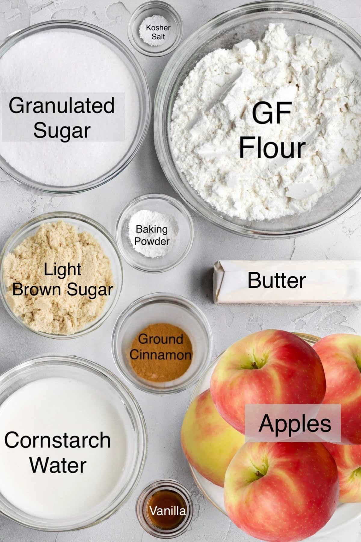 Easy apple cobbler ingredients in separate bowls: granulated sugar, kosher salt, gluten free flour, baking powder, light brown sugar, butter, ground cinnamon, cornstarch water, apples, and vanilla.